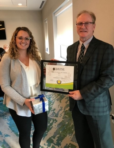 White man wearing glasses and a suit accepts a framed award from a woman. 