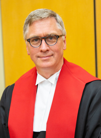 White man with black rimmed glasses and wearing black, red and white judicial robes.