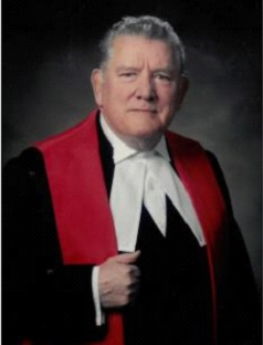 Grey-haired man wearing red, white and black judicial robes. 