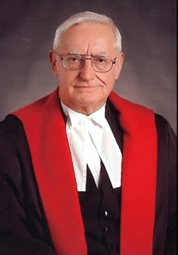 White man with white hair and glasses wearing black, red and white judicial robes. 