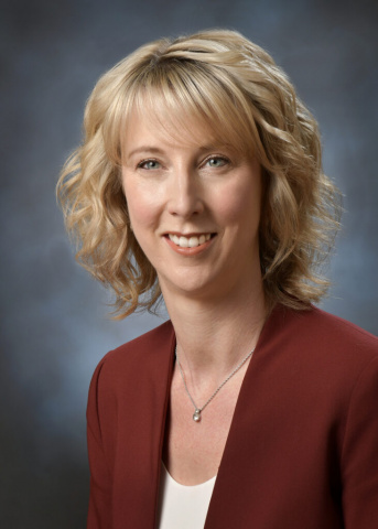 White woman with short wavy blonde hair wearing a red blazer over a white shirt.