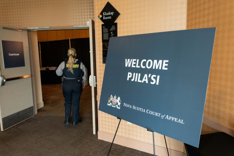 A photo of a sign on an easel with the words "Welcome. Pjila'si." In the background is Sheriff standing next to a door with a sign that says "Courtroom."