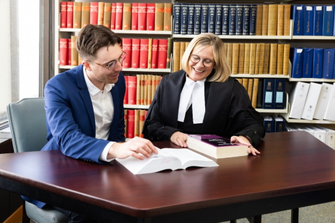 A female judge in black robes and a man in a blue suit sit at a desk in front of an open laptop. Behind them are shelves of legal books. 