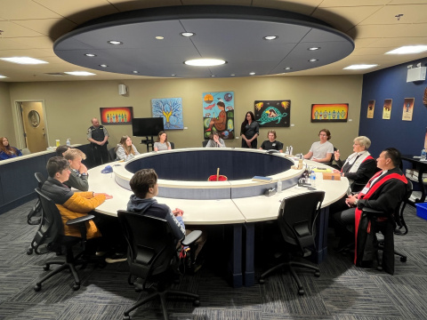 Judges in robes and students sit around a circular table in a courtroom with Indigenous artwork on the walls. 