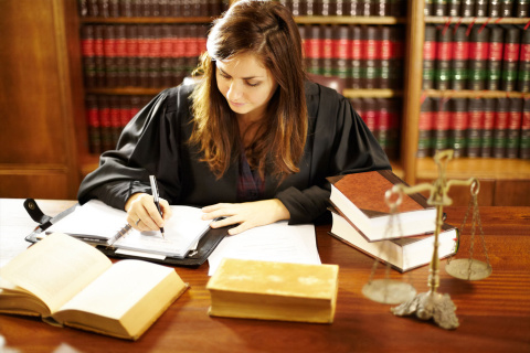 Young woman with long brown hair seated at a desk, writing in a notebook with textbooks open in front of her.