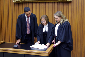 Young male and female law clerks review legal papers with a female judge. They are all wearing black robes.  