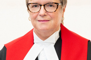 White woman with glasses and short blonde hair wearing red, black and white judicial robes. 