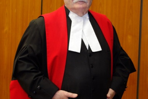 White man with glasses wear black, red and white judicial robes and his hands in his pockets.