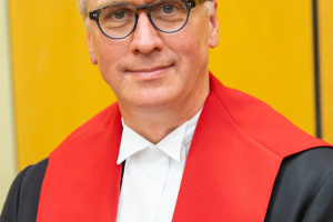 White man with black rimmed glasses and wearing black, red and white judicial robes.