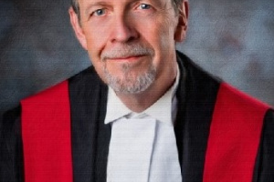 White man with a goatee wearing red, black and white judicial robes. 