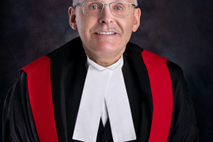 Smiling white man with glasses wearing red, black and white judicial robes. 