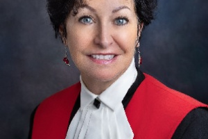Female judge with short dark hair, wearing black and red judicial robes with a lacy collar. 