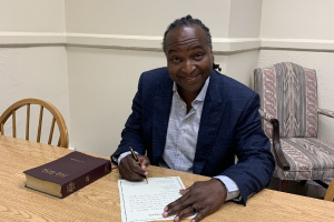 Black man in a suit jacket seated at a table signing a document. 
