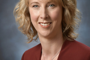 White woman with short wavy blonde hair wearing a red blazer over a white shirt.